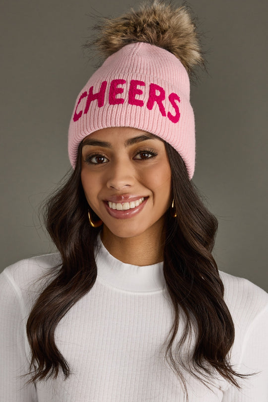 Woman wearing a pink 'CHEERS' beanie with a fur pom-pom on a gray background
