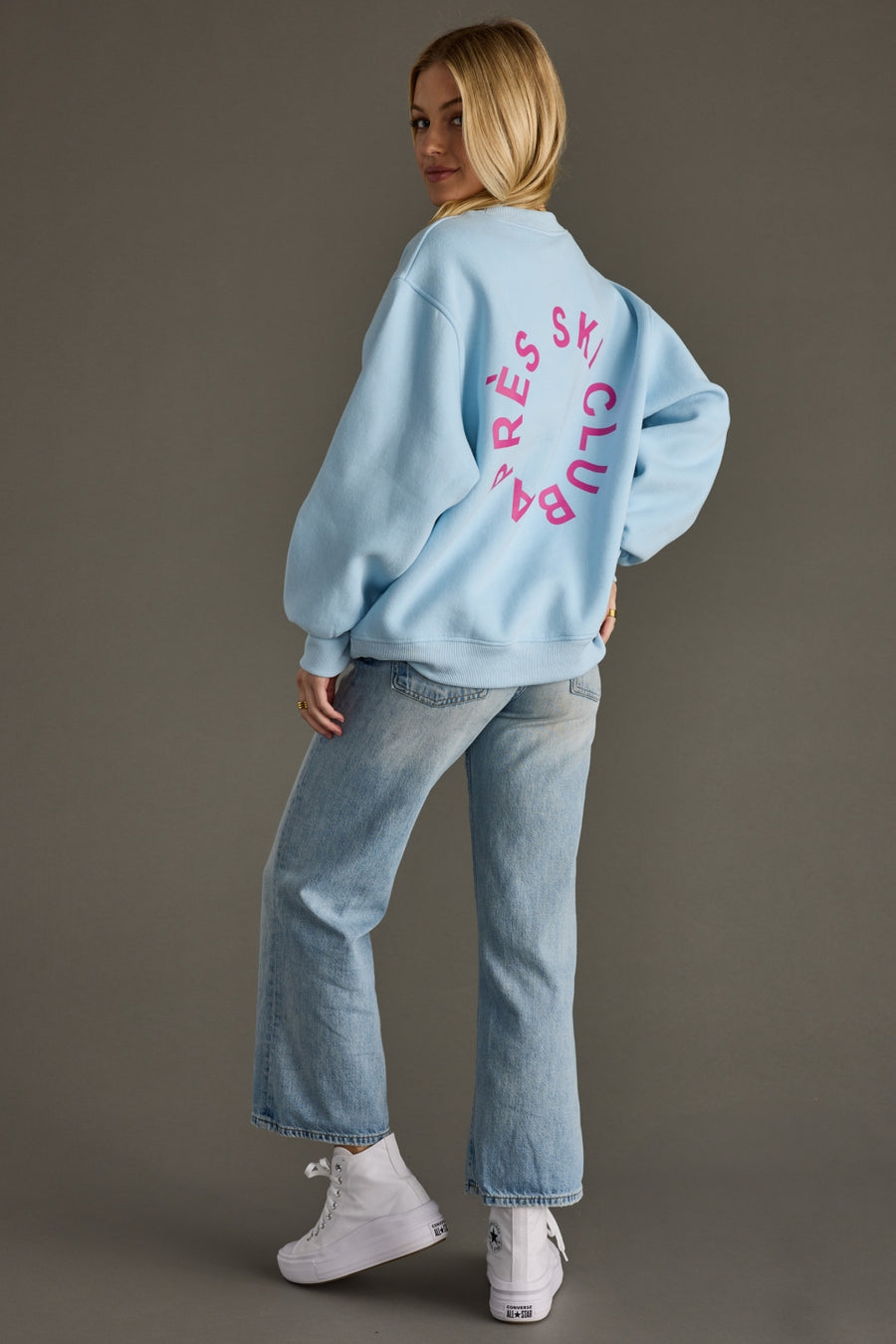 Person wearing a light blue sweatshirt with pink text on the back, standing against a gray background