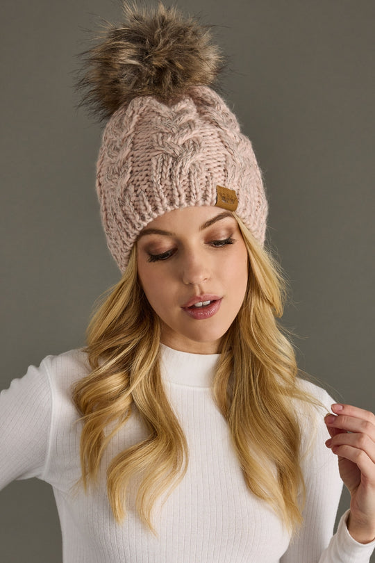 A woman with long blonde hair wears the Panache Apparel Collection Heartbeat Pom Hat, a light pink fleece-lined beanie with a faux fur pom, and a white ribbed turtleneck, looking down with a neutral expression against a gray background.
