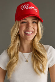 A smiling woman with long blonde hair wears the Panache Apparel Collection Merry Trucker Hat in red & white, a white short-sleeve top, and a gold necklace, standing against a gray background.