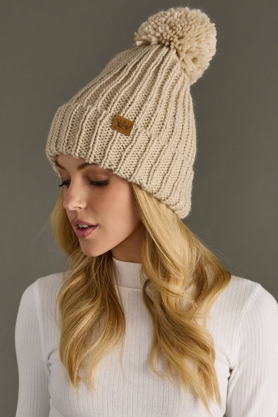 Woman wearing a beige knitted beanie with a pom-pom against a gray background