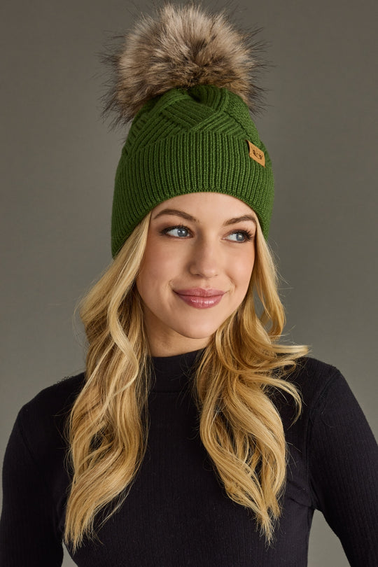 Woman wearing a green knit beanie with a fur pom-pom against a gray background