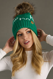 Person wearing a green knit beanie with a fur pom-pom on a gray background