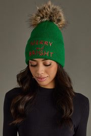 Green knit beanie with fur pom-pom and 'Merry and Bright' text worn by a model against a gray background