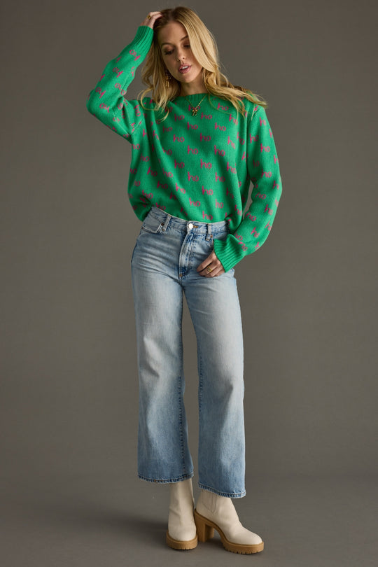 Woman wearing a green sweater with pink patterns, light blue jeans, and white boots on a gray background
