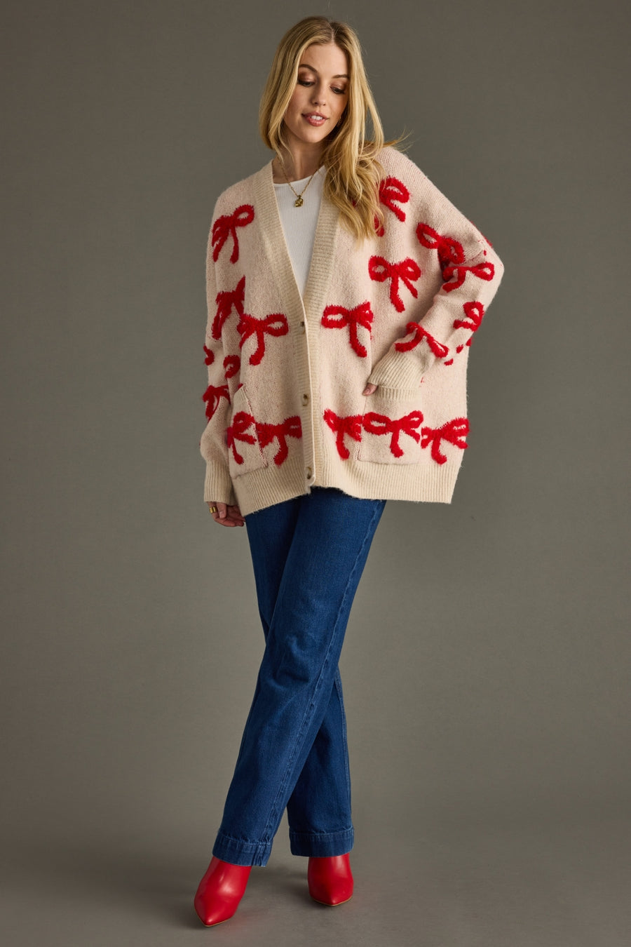 Woman wearing a cream cardigan with red bow patterns, blue jeans, and red shoes on a gray background