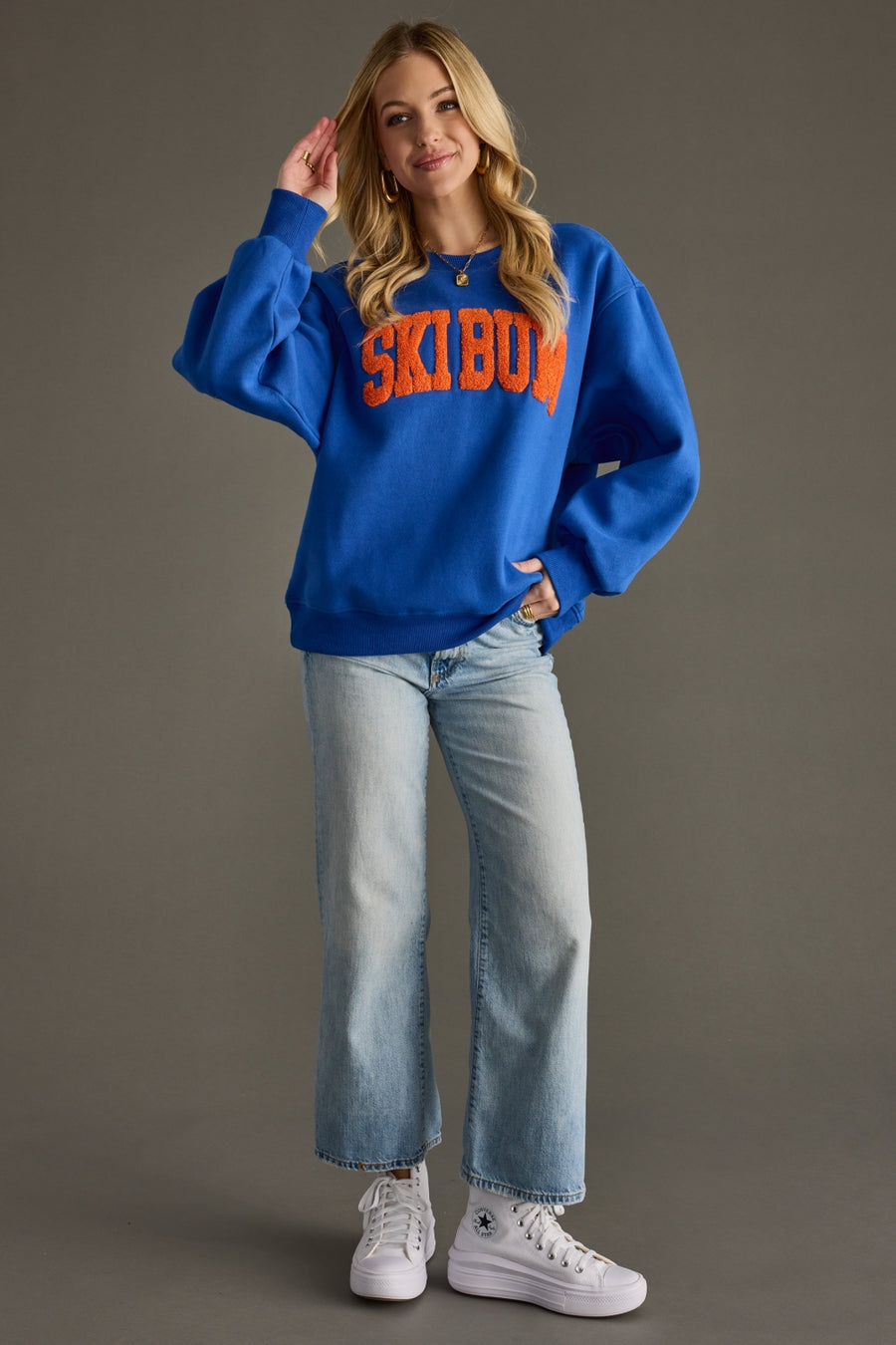 Person wearing a blue sweatshirt with 'Ski Bum' text, light blue jeans, and white sneakers on a gray background