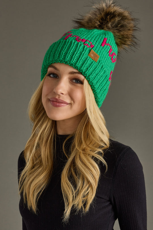 Woman wearing a green knit beanie with a fur pom-pom against a gray background