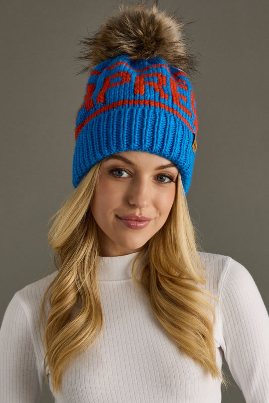 Woman wearing a blue beanie with orange text and a fur pom-pom on a gray background