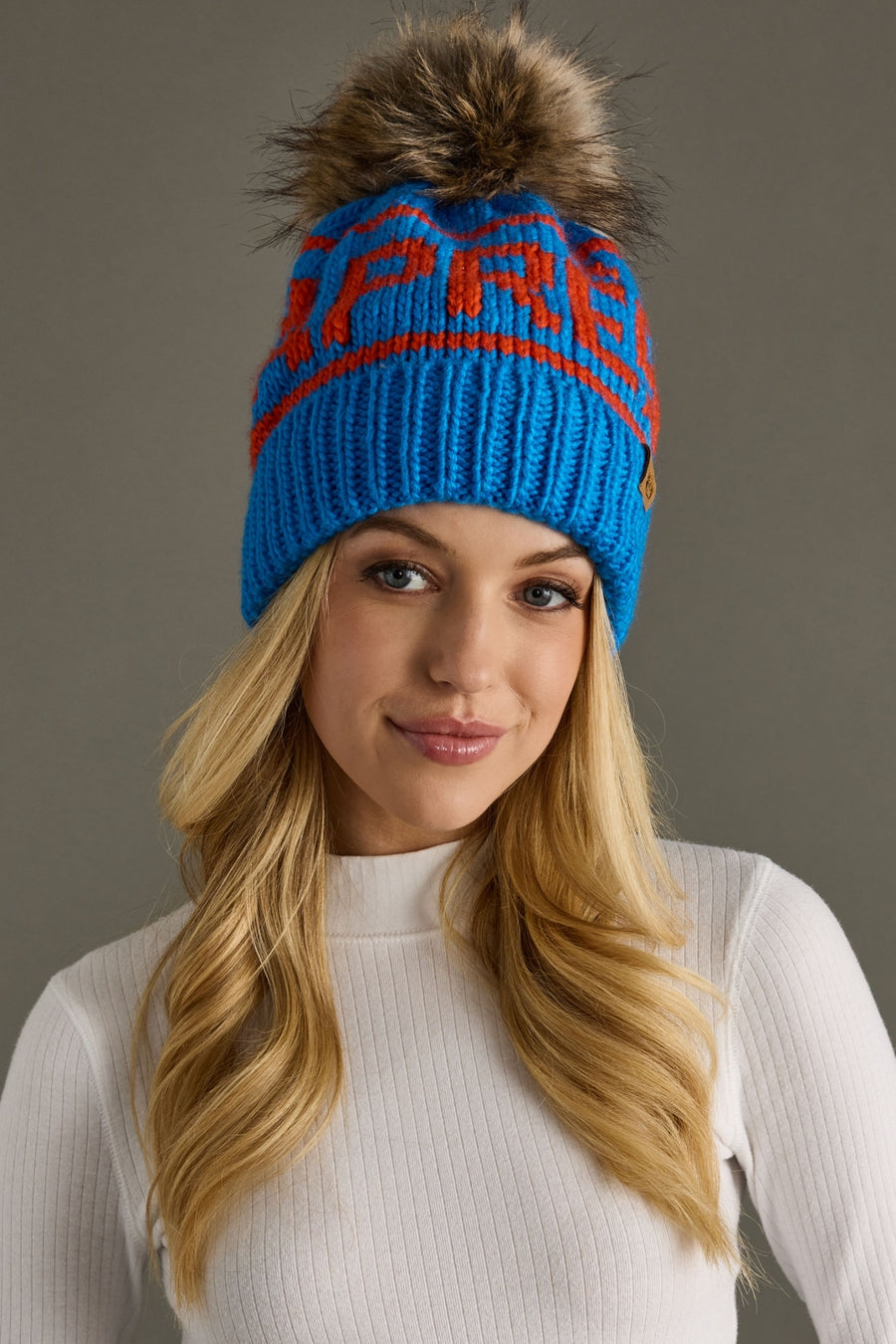 Woman wearing a blue beanie with orange text and a fur pom-pom on a gray background