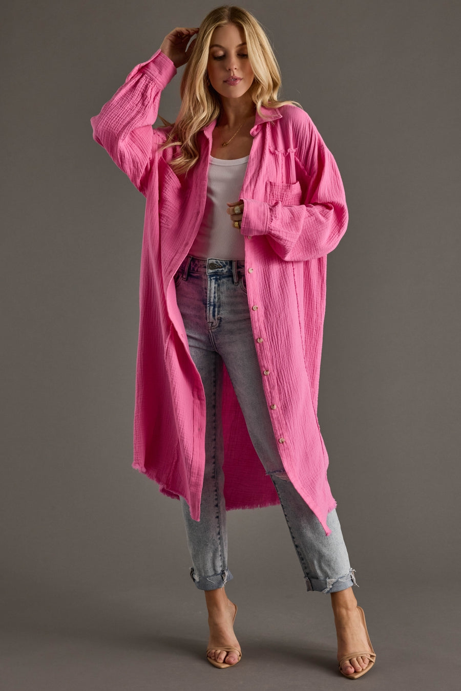 Woman wearing a long pink button up over a white top and jeans on a gray background
