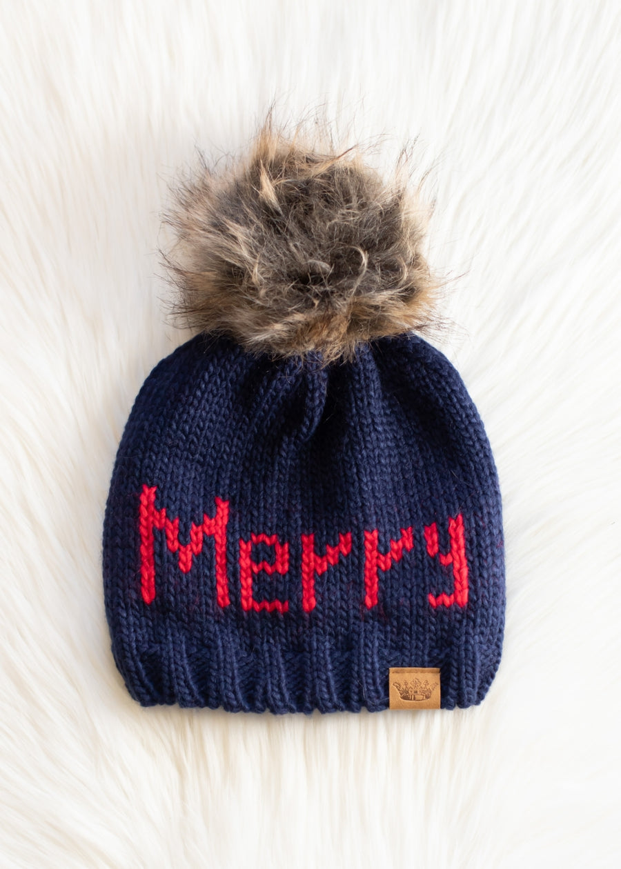 Navy blue knit beanie with red 'Merry' text and fur pom-pom on a white background