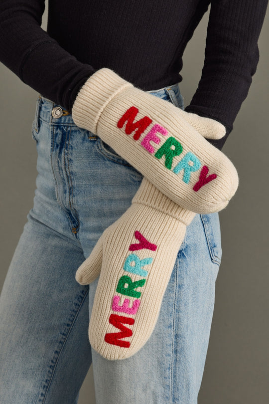 Beige mittens with colorful 'MERRY' text worn by a person in jeans and a black sweater.