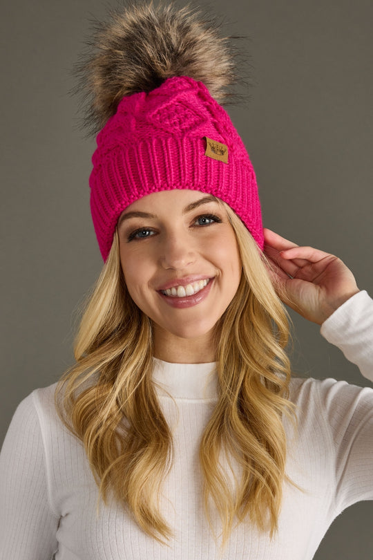 Woman wearing a bright pink beanie with a fur pom-pom against a gray background