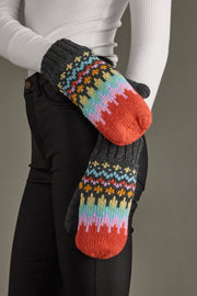 Colorful knitted mittens with a pattern held by a person wearing a white sweater and black pants.