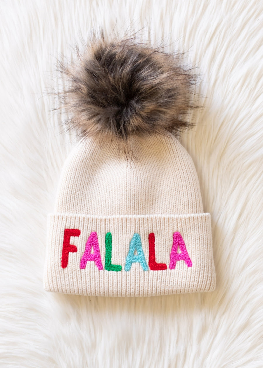 The Fa La La Pom Hat by Panache Apparel Collection is a cream knit pom hat, fleece lined for warmth, featuring colorful FALALA lettering on the brim and topped with a large faux fur pom-pom, shown on a white fluffy surface.