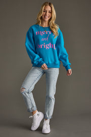 A smiling woman poses on one leg in the Merry & Bright Sweatshirt - Blue by Panache Apparel Collection, paired with ripped light blue jeans and white sneakers, set against a plain gray background.