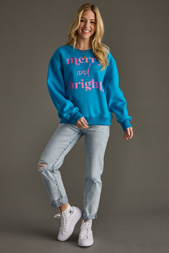 A smiling woman poses on one leg in the Merry & Bright Sweatshirt - Blue by Panache Apparel Collection, paired with ripped light blue jeans and white sneakers, set against a plain gray background.