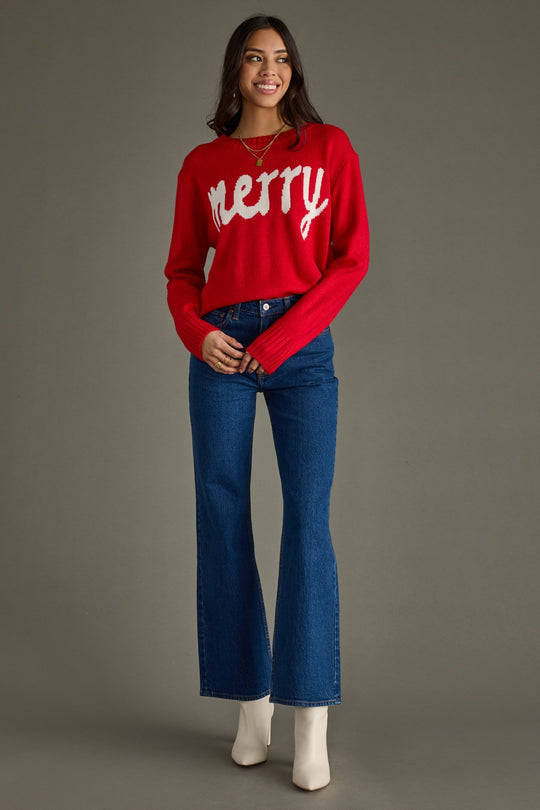Woman wearing a red 'Merry' sweater and blue jeans on a gray background