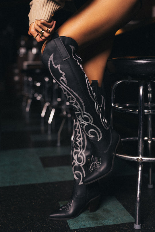 A person wearing Panache Apparel Collection’s Black Embroidered Western Boots sits on a bar stool with one leg crossed over the other. In the background, additional stools and a dark, patterned floor are visible.