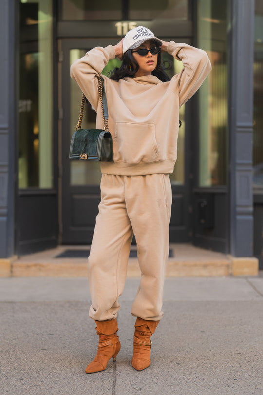 A person wearing a khaki hoodie and cozy Khaki Panache Sweatpants, a baseball cap, dark sunglasses, and heeled brown boots, carrying a dark green handbag with a chain strap.