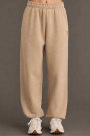 Model showcases the Khaki Panache Sweatpants from Panache Apparel Collection, featuring a beige fleece fabric and elastic waistband. Styled with white chunky sneakers, the relaxed-fit design emphasizes comfort and contemporary flair, photographed against a plain grey background with only the lower body in view.