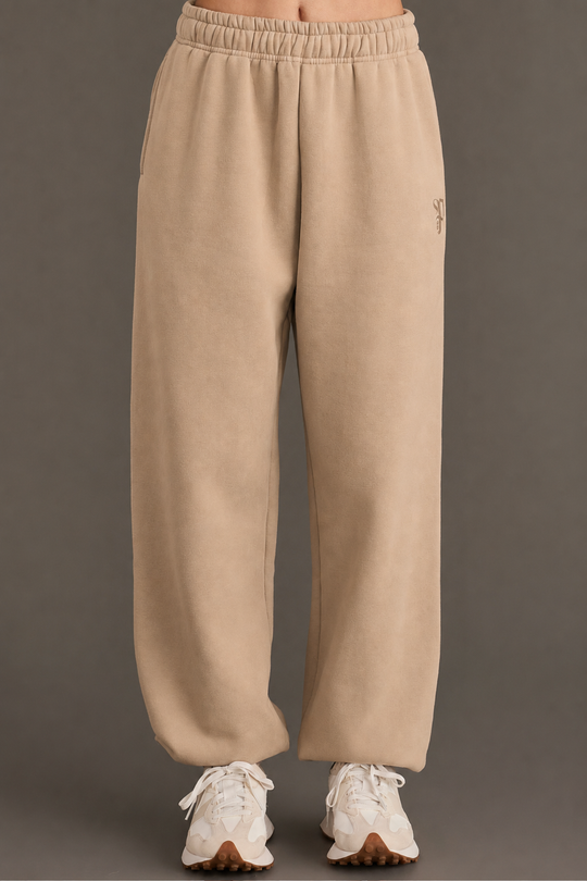 Model showcases the Khaki Panache Sweatpants from Panache Apparel Collection, featuring a beige fleece fabric and elastic waistband. Styled with white chunky sneakers, the relaxed-fit design emphasizes comfort and contemporary flair, photographed against a plain grey background with only the lower body in view.