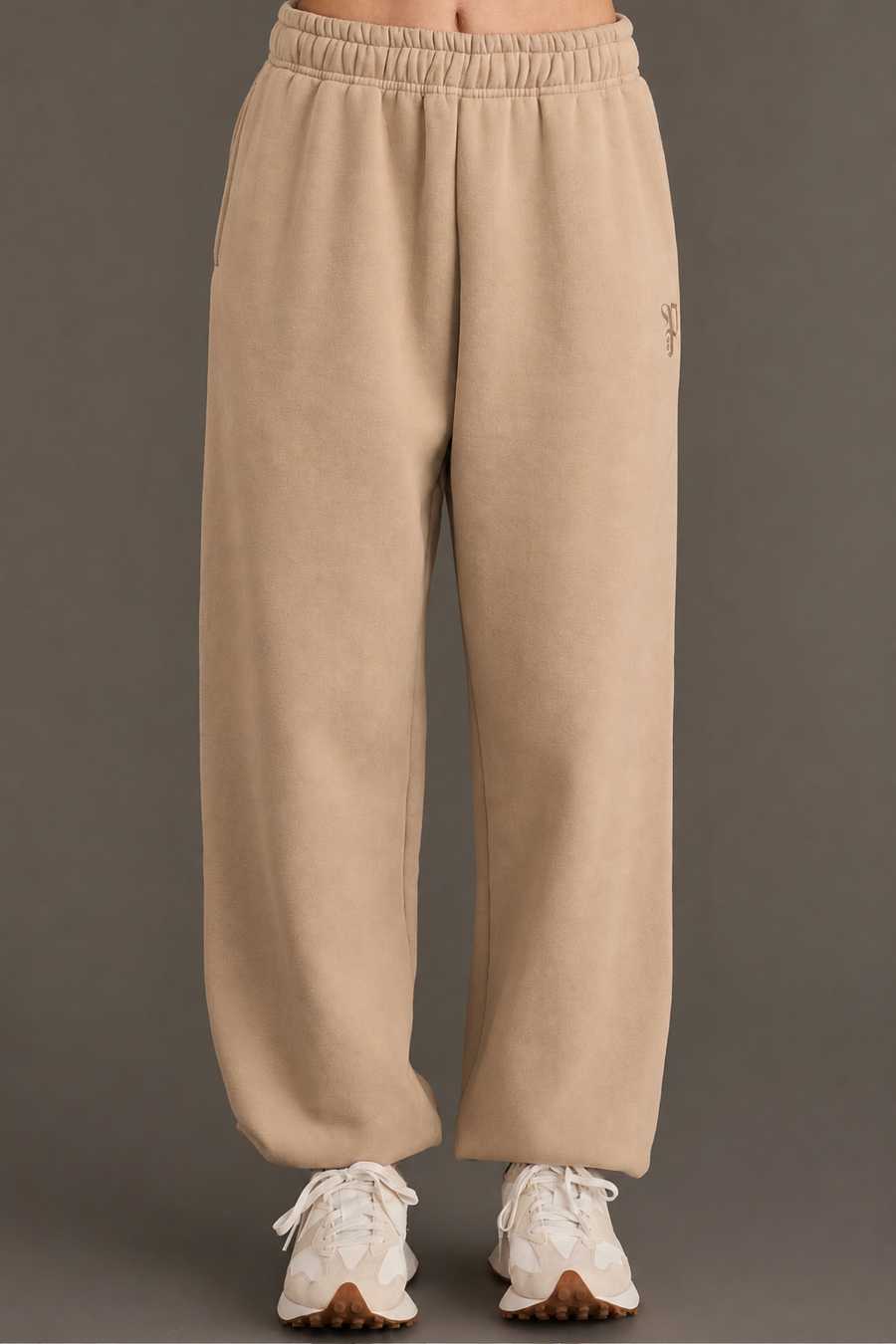 Model showcases the Khaki Panache Sweatpants from Panache Apparel Collection, featuring a beige fleece fabric and elastic waistband. Styled with white chunky sneakers, the relaxed-fit design emphasizes comfort and contemporary flair, photographed against a plain grey background with only the lower body in view.