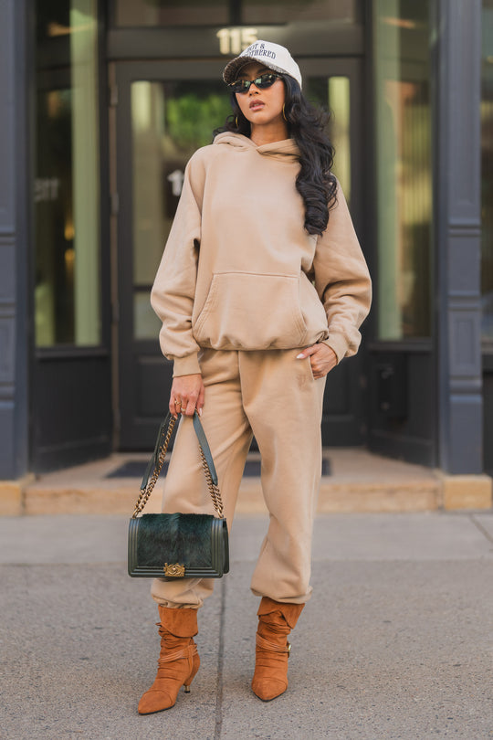 A woman in a tan hoodie and matching Khaki Panache Sweatpants wears a baseball cap, sunglasses, and tan ankle boots, carrying a dark green handbag.