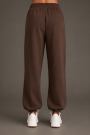 A person stands facing away, wearing Panache Apparel Collection’s Brown Panache Sweatpants—relaxed fit with elastic cuffs—paired with white sneakers against a plain background.