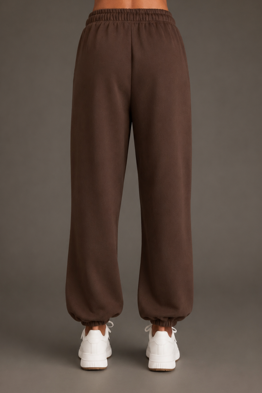 A person stands facing away, wearing Panache Apparel Collection’s Brown Panache Sweatpants—relaxed fit with elastic cuffs—paired with white sneakers against a plain background.