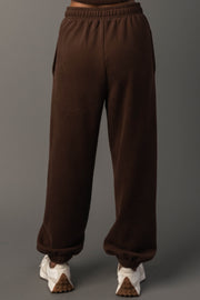 A person stands facing away, wearing the Brown Panache Sweatpants from Panache Apparel Collection and white sneakers, against a plain gray background.