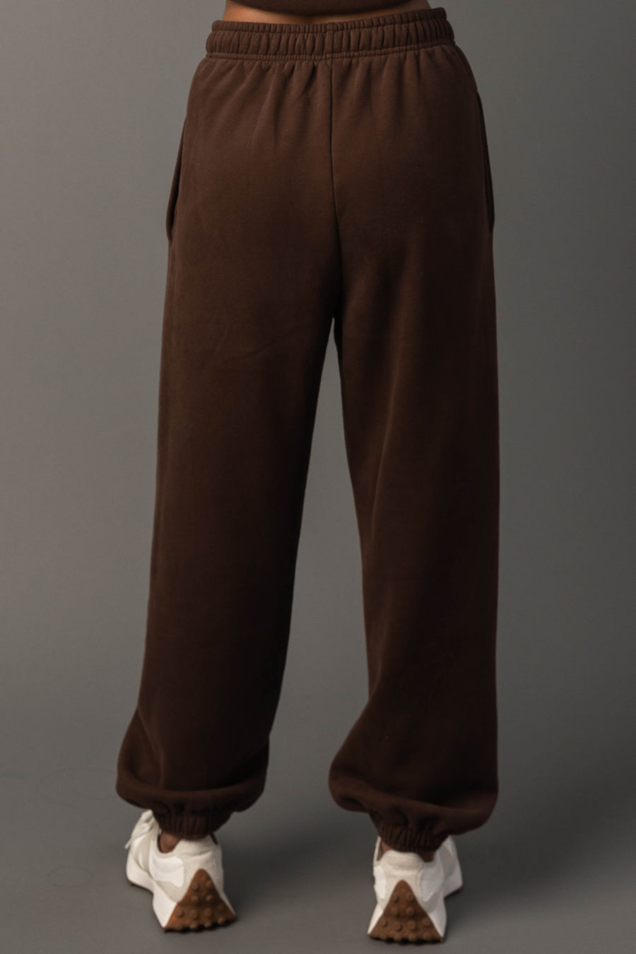 A person stands facing away, wearing the Brown Panache Sweatpants from Panache Apparel Collection and white sneakers, against a plain gray background.