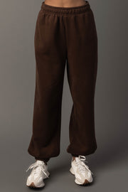 A person wearing Panache Apparel Collection’s Brown Panache Sweatpants and white sneakers stands against a plain gray background.