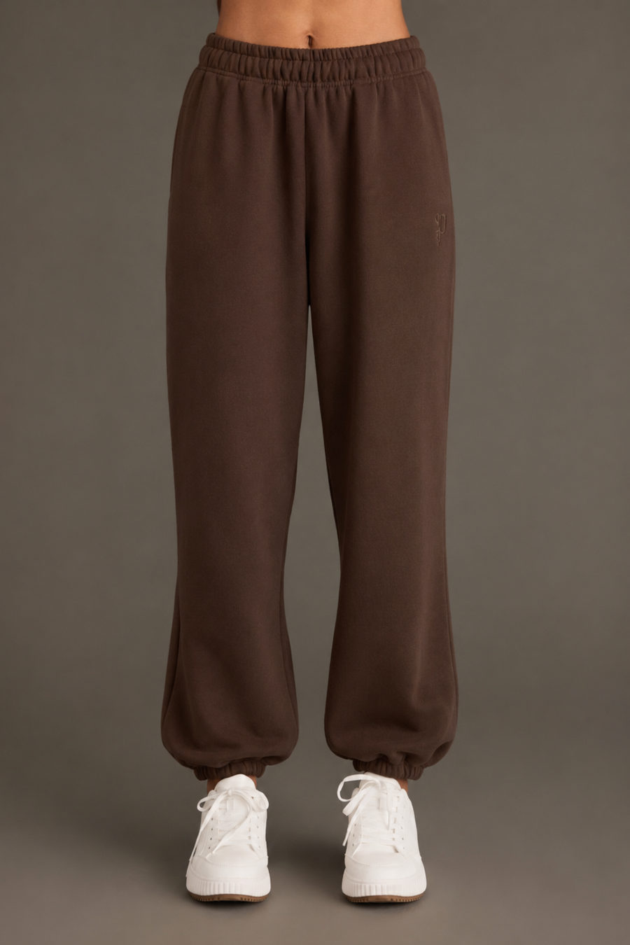 A person models the Brown Panache Sweatpants from the Panache Apparel Collection, paired with white sneakers and photographed against a plain gray background. The image is cropped at the waist to highlight the relaxed fit and warm brown hue of the sweatpants.