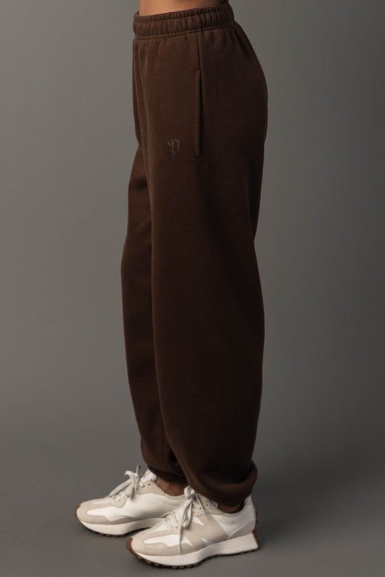 A person wearing the Brown Panache Sweatpants from the Panache Apparel Collection and white sneakers stands against a plain gray background, with only the lower half of their body visible in the image.