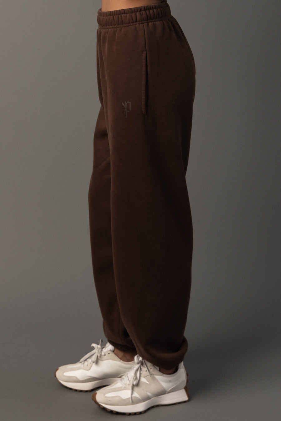 A person wearing the Brown Panache Sweatpants from the Panache Apparel Collection and white sneakers stands against a plain gray background, with only the lower half of their body visible in the image.