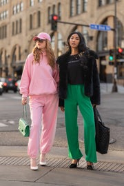 Two women stand on a city street corner: one wearing a pink hooded sweatshirt and Light Pink Panache Sweatpants with a green handbag, the other wearing a black jacket and bright green trousers with a black handbag.