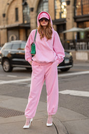 A person in a matching light pink hooded sweatshirt and cozy Light Pink Panache Sweatpants, wearing a pink baseball cap, sunglasses, and white high heels while carrying a small green handbag.