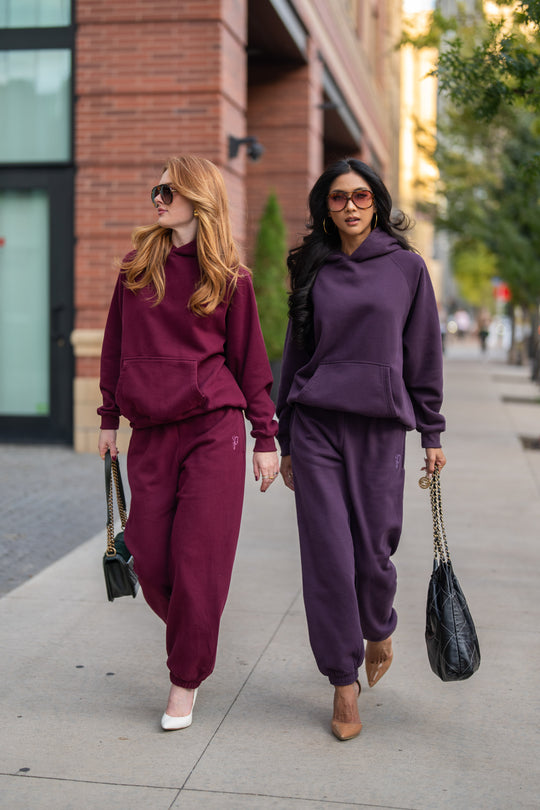 Two women walk on a sidewalk wearing matching hooded sweatshirts and Plum Panache Sweatpants, one in deep red and the other in dark purple, both carrying shoulder bags.