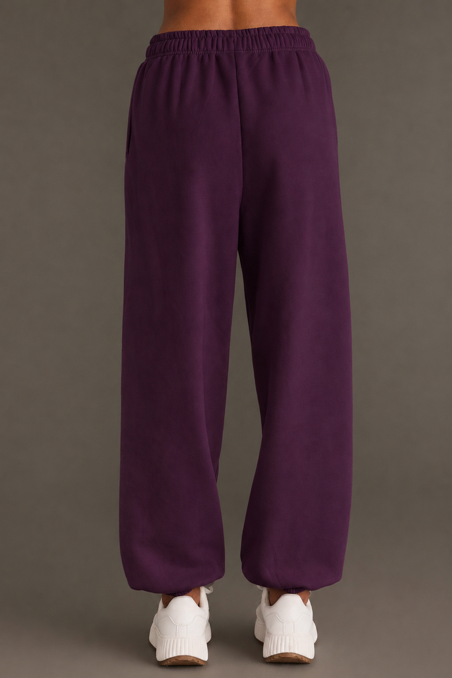 A person stands facing away, wearing loose-fitting Panache Apparel Collection Plum Panache Sweatpants and white sneakers, against a plain gray background.
