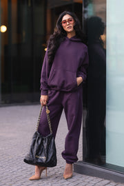 A person wears a dark purple hooded sweatshirt and matching Plum Panache Sweatpants, tan high heels, and a black quilted leather handbag.