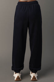 A person wearing Dark Navy Panache Sweatpants from Panache Apparel Collection and white sneakers stands against a plain gray background. The image is taken from behind, showcasing the back view of the sweatpants and shoes.