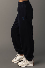 A person wearing Panache Apparel Collection’s Dark Navy Panache Sweatpants and white sneakers stands on a gray surface, with their side facing the camera. The sweatpants feature a small blue logo on the upper thigh.