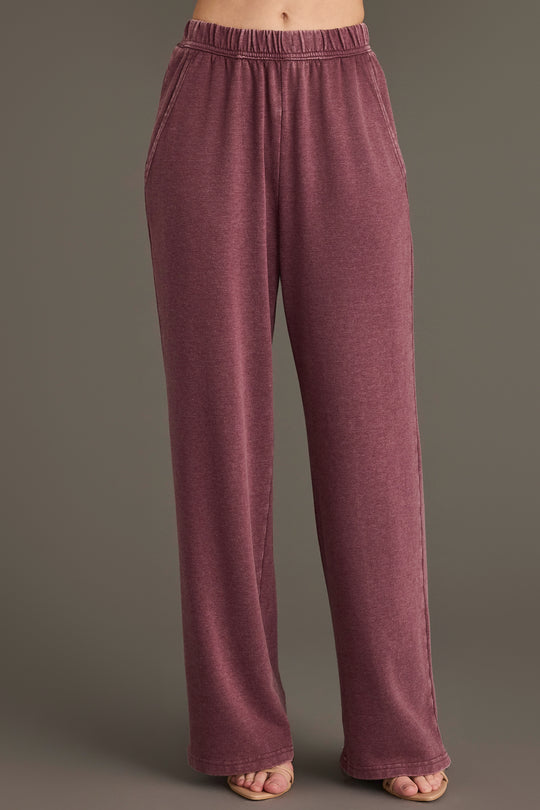 A person wearing the Panache Apparel Collection Merlot Washed Soft Sweatpants, which are high-waisted, loose-fitting, and feature pockets in a mauve hue, stands against a plain background with only their lower torso and legs visible.