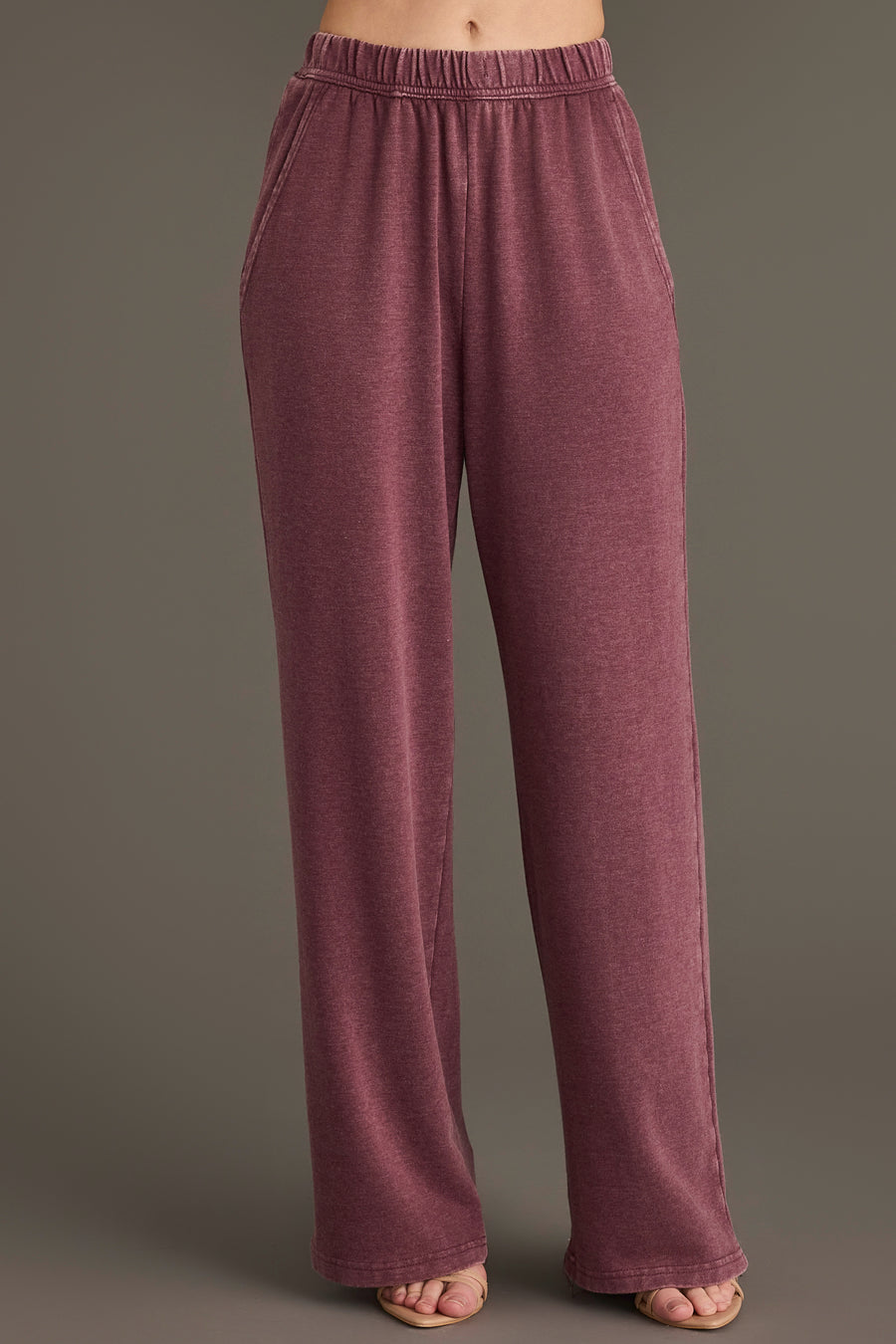 A person wearing the Panache Apparel Collection Merlot Washed Soft Sweatpants, which are high-waisted, loose-fitting, and feature pockets in a mauve hue, stands against a plain background with only their lower torso and legs visible.