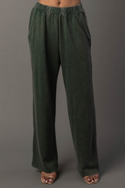 A person wearing Panache Apparel Collection’s Pine Green Washed Soft Sweatpants, featuring a relaxed fit, elastic waistband, and side pockets, stands against a plain gray background. Their feet are shown in open-toe sandals.