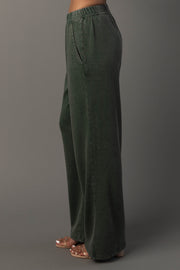 A person is shown from the side against a plain gray background, wearing the Pine Green Washed Soft Sweatpants from Panache Apparel Collection. The wide-leg sweatpants feature an elastic waistband and pockets, and the model’s toes are visible in clear sandals.