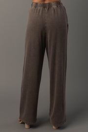 A person models the Cocoa Washed Soft Sweatpants from Panache Apparel Collection. A loose-fitting, wide-leg brown pants with an elastic waistband, and standing against a plain gray background. The back view highlights the fit and drape of the sweatpants.