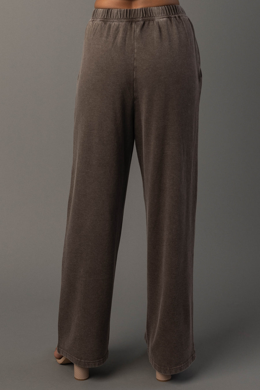 A person models the Cocoa Washed Soft Sweatpants from Panache Apparel Collection. A loose-fitting, wide-leg brown pants with an elastic waistband, and standing against a plain gray background. The back view highlights the fit and drape of the sweatpants.